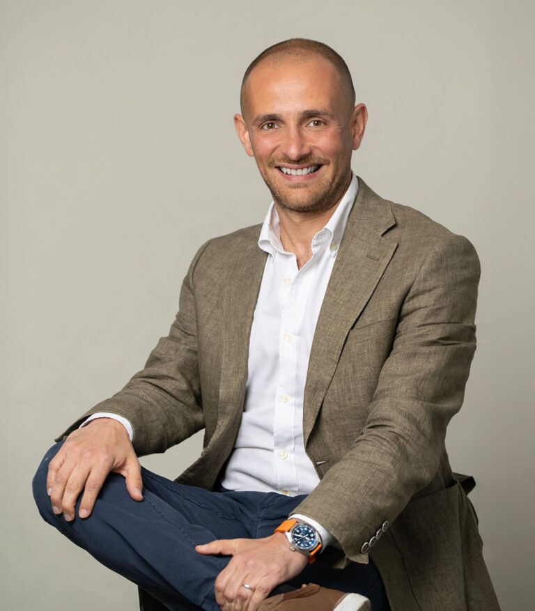 Smiling man in a suit sitting casually with arms crossed, against a neutral background.