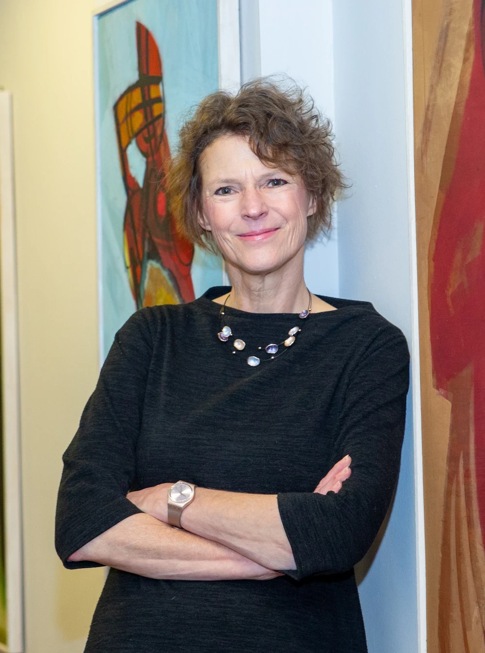 Professional woman smiling with arms crossed in an art gallery setting, surrounded by colorful artwork.