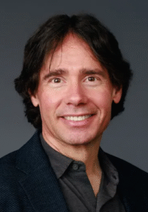 Professional man smiling, wearing a dark jacket, with shoulder-length hair and a neutral background.