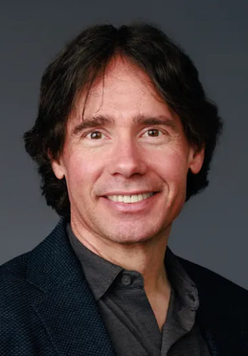 Professional man smiling, wearing a dark jacket, with shoulder-length hair and a neutral background.