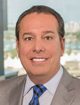 Male attorney smiling, wearing a gray suit and blue tie, with a blurred cityscape in the background.