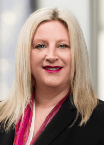 Lawyer with long blonde hair and a pink scarf, smiling in a professional office setting.