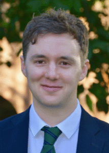 Professional man in suit and tie smiling outdoors, trees visible in the background.