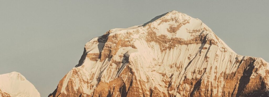 Snow-capped mountain peak under clear sky with rugged terrain in foreground.