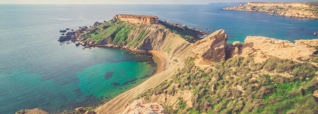 Coastal landscape featuring rocky cliffs, clear blue waters, and lush green hills under a clear sky.