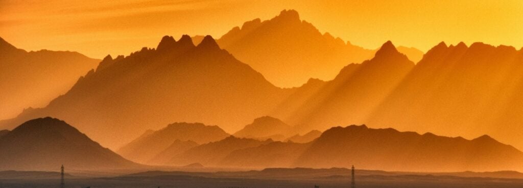 Sunset over mountainous landscape with silhouettes and gradient orange sky, creating a serene natural scene.
