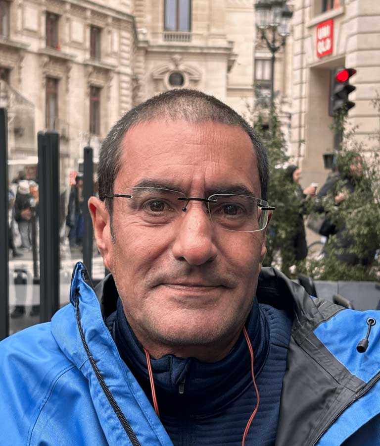 Man with glasses wearing a blue jacket, seated outdoors in an urban setting with people in the background.