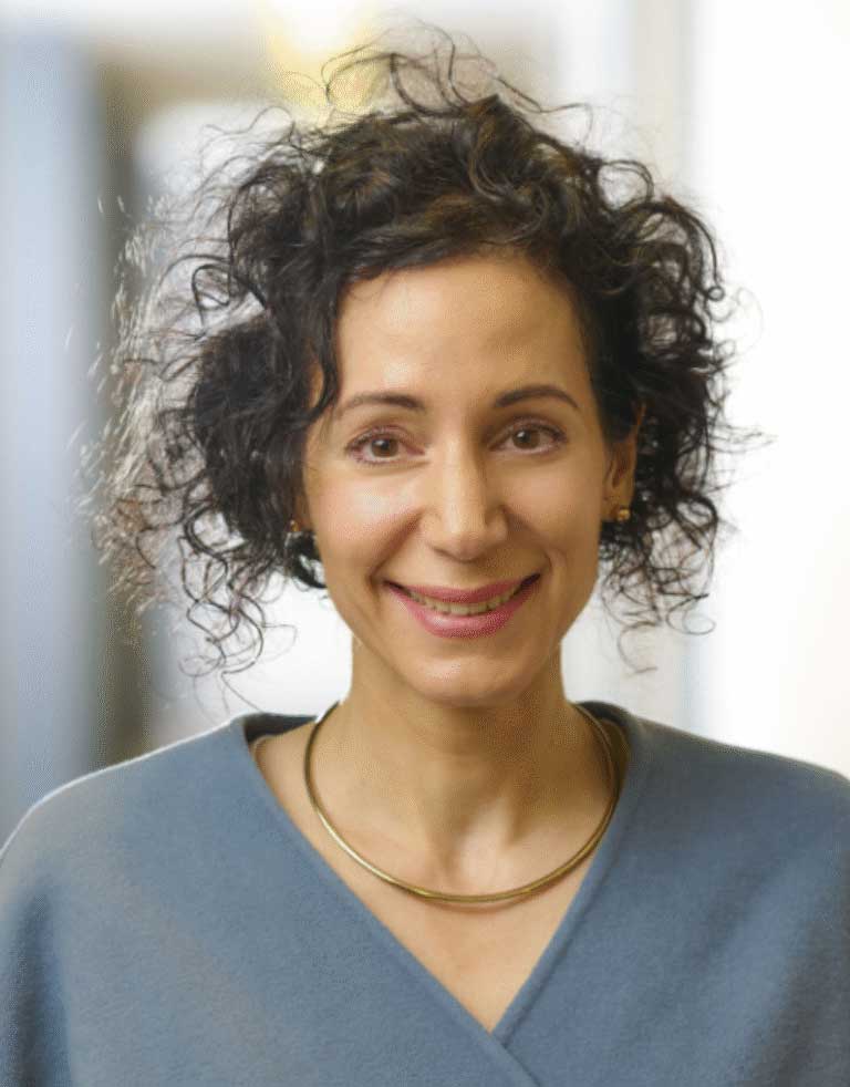 Woman with curly hair smiling, wearing a gray top and a gold necklace, in a professional setting.