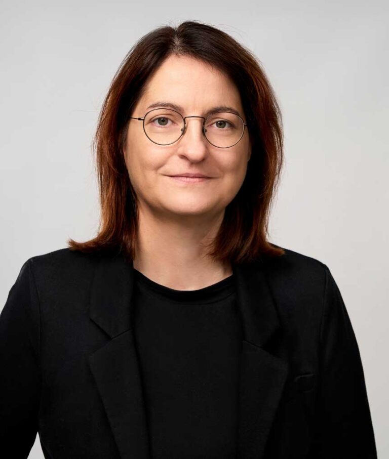 Legal professional with shoulder-length hair, wearing a black blazer and glasses, posing against a neutral background.