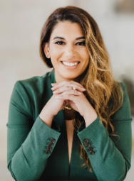 Attorney smiling with hands clasped, wearing a green blazer, seated in a professional environment.