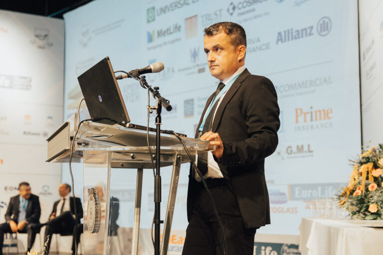 Speaker presenting at a conference with a laptop and microphone, featuring company logos in the background.