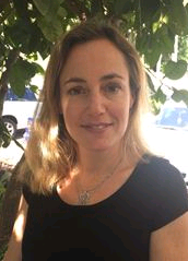 Professional woman with light hair standing outdoors in front of leafy greenery.