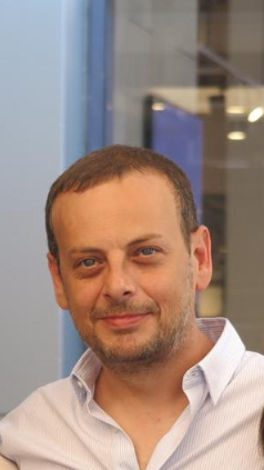 Bearded man in a casual shirt smiling in an outdoor urban setting.