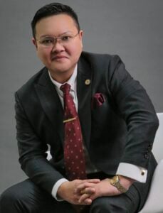 Lawyer in a black suit with glasses, seated and looking at the camera in a professional setting.