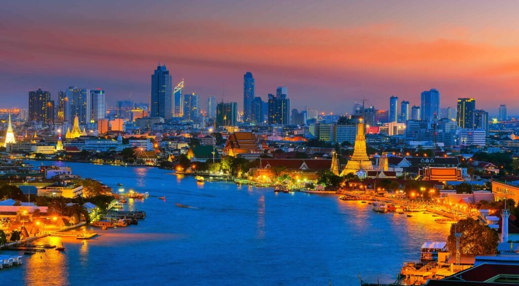 City skyline at dusk with lit buildings and temples along the river, reflecting vibrant colors in the water.