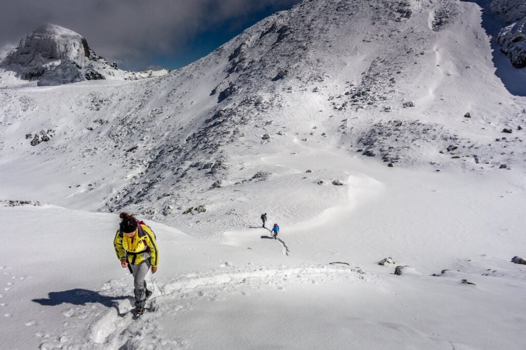 Three hikers ascending a snowy mountain trail surrounded by rocky terrain and blue skies.