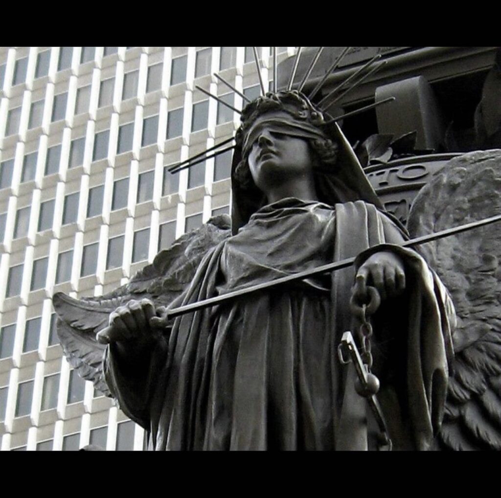 Statue of Lady Justice with blindfold, holding scales and chains, set against a modern building backdrop.