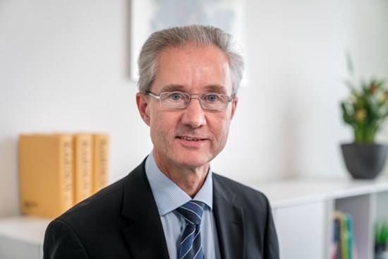 Lawyer In Professional Attire Posing For A Portrait In An Office Setting With Legal Documents In The Background.