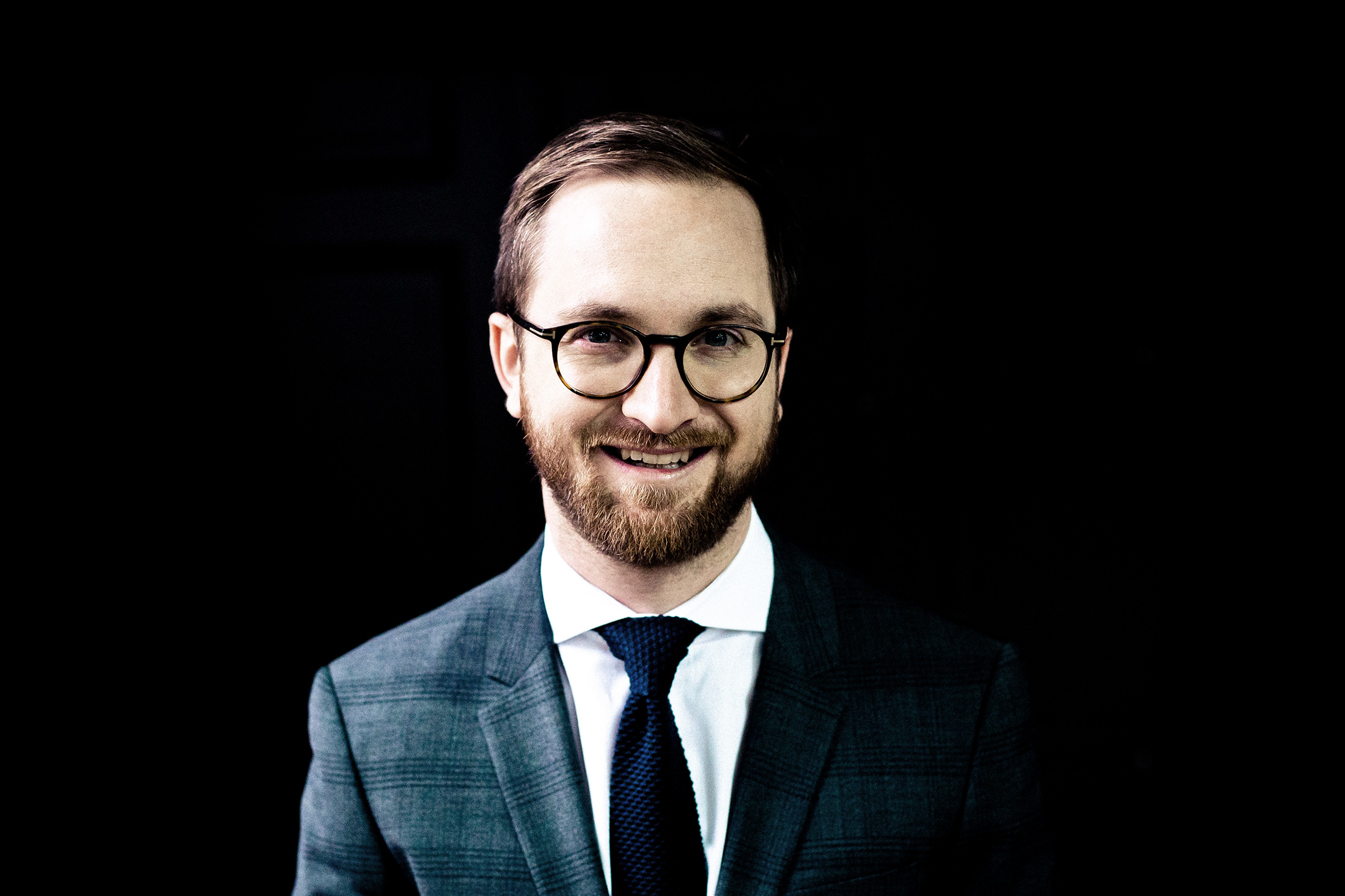 Lawyer smiling while wearing a suit and glasses against a dark background.