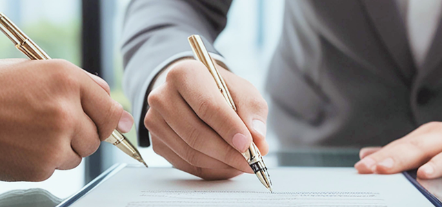 Two individuals signing legal documents, each holding a pen in a professional setting.