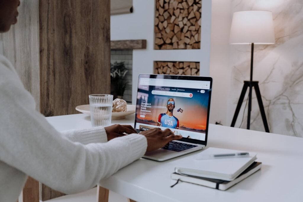 Person using a laptop at a table, focused on an online platform, with a glass of water and a notebook nearby.
